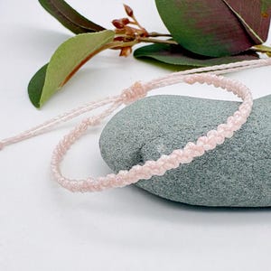 May include: A light pink macrame bracelet with an adjustable sliding knot closure. The bracelet is displayed on a grey stone, with green leaves and brown stems in the background. The bracelet is made of a woven, textured material.