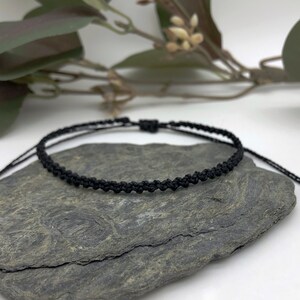 Puede incluir: Un brazalete ajustable trenzado negro con un dise&ntilde;o simple. El brazalete est&aacute; hecho de cord&oacute;n delgado y est&aacute; atado con un nudo en el extremo.