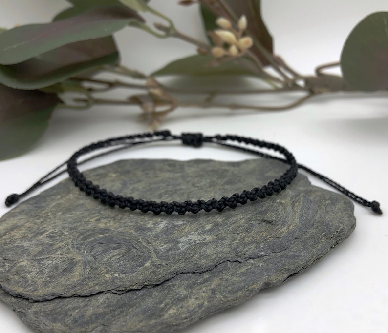 Puede incluir: Un brazalete ajustable trenzado negro con un dise&ntilde;o simple. El brazalete est&aacute; hecho de cord&oacute;n delgado y est&aacute; atado con un nudo en el extremo.