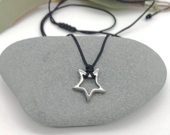 May include: A silver-colored star pendant necklace with a black cord. The star is a five-pointed shape with a hollow center. The necklace is displayed on a gray stone, with a white background.