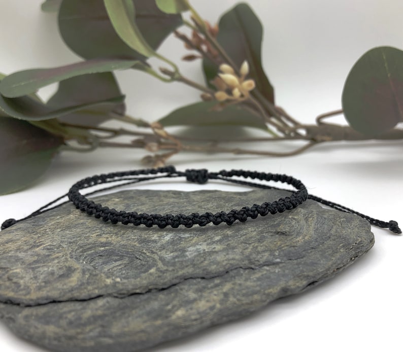 Puede incluir: Un brazalete trenzado negro con un dise&ntilde;o anudado. El brazalete est&aacute; sobre una roca gris.