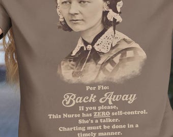 May include: Brown t-shirt featuring a sepia-toned portrait of Florence Nightingale with text that reads "Back Away" and other humorous phrases. The shirt is a solid brown color.