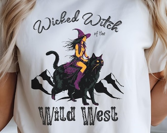 May include: Cream-colored t-shirt featuring a graphic of a witch riding a black cat, with the text "Wicked Witch of the Wild West." The witch is wearing a purple outfit and hat. Mountains are in the background.