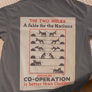 Può includere: T-shirt grigia con grafica in stile vintage. La grafica include il testo "THE TWO MULES" e "A fable for the Nations". Mostra illustrazioni di muli, con la morale "CO-OPERATION is better than Conflict".