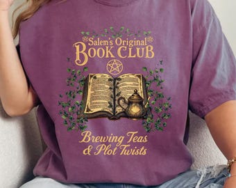 May include: A purple t-shirt with the text "Salem's Original Book Club" above an illustration of an open book, teapot, and foliage. The phrase "Brewing Teas & Plot Twists" is below the illustration. The shirt is worn by a person.