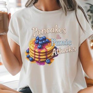 Könnte beinhalten: Weißes T-Shirt mit einem Grafikdruck von Pfannkuchen und Blaubeeren und dem Text "Pancakes & Panic Attacks".