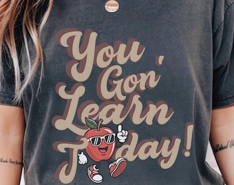 May include: Dark gray t-shirt with the text "You Gon' Learn Today!" in brown and tan lettering. The shirt features a cartoon apple wearing sunglasses and sneakers, giving a thumbs up.