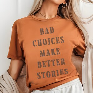 May include: A rust-colored t-shirt with the text "BAD CHOICES MAKE BETTER STORIES" in a distressed, dark brown font. The shirt has short sleeves and a relaxed fit. The model is wearing a gold necklace and hoop earrings.