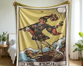 May include: A large, rectangular blanket featuring a tarot card design. The card depicts a figure in colorful clothing, a dog, and the sun. The text "THE FOOL" is at the bottom. The blanket has a yellow background with a green border.