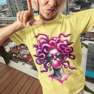 May include: Yellow t-shirt featuring a graphic of Medusa with pink snakes, sunglasses, and a bubblegum bubble. The artwork has a watercolor style with pink and purple accents. The shirt is a casual, short-sleeved style.