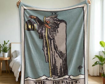 May include: A rectangular, light blue blanket featuring a tarot card design. The image depicts a robed figure holding a lantern and staff, with the words "THE HERMIT" at the bottom. The blanket has a white border and a cream-colored frame.