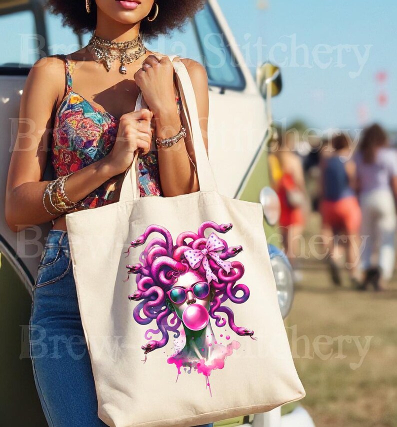 May include: A cream-colored tote bag featuring a vibrant illustration of Medusa with pink hair, sunglasses, and a bubblegum bubble. The Medusa graphic is surrounded by pink snakes and a pink bow. The bag is held by a person wearing a colorful top and blue jeans.
