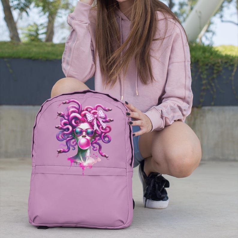 May include: A light purple backpack featuring a Medusa-inspired graphic. The illustration shows Medusa with pink hair, sunglasses, and a bubblegum bubble. The backpack has a front pocket and adjustable straps, perfect for school or travel.