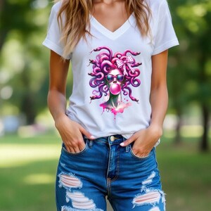 May include: White V-neck t-shirt featuring a graphic of Medusa with pink hair, sunglasses, and a bubblegum bubble. The design includes a pink bow and snakes. The model is wearing blue denim shorts.