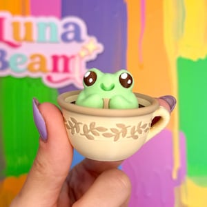 May include: A light green frog figurine in a small beige teacup with floral detailing. The teacup is held in a hand with purple nail polish. The background is a colorful abstract design with the text "Luna Beam".