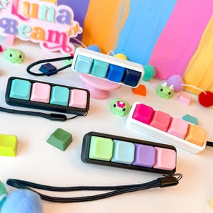 May include: A selection of keycap accessories in pastel colours, including sets of four keycaps. The image shows a "Luna Beam" logo and decorative pom-poms against a vibrant, abstract painted background.