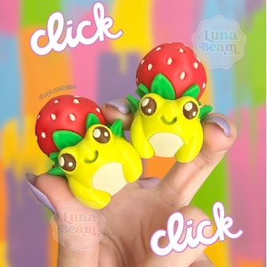 May include: Two yellow frog figurines with strawberry hats are held up. The frogs have big eyes and are smiling. The strawberries are red with green leaves. The word "click" is written in a cursive font in two places.