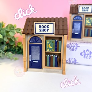 May include: Miniature book shop model with a brown tiled roof, a blue door, and a sign that reads "BOOK SHOP". The shop has a shelf with books and a crescent moon design. The word "click" is written in a cursive font.