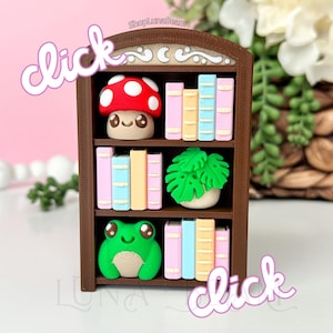May include: A miniature brown bookshelf with colorful books, a red mushroom, a green frog, and a green plant. The word "click" is written in a cursive font on the left and right sides of the bookshelf.