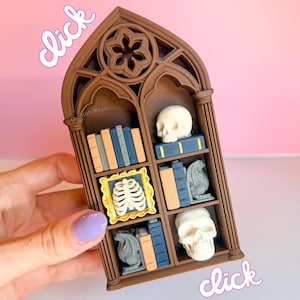 May include: A miniature brown bookshelf with gothic arch design, filled with tiny books, skulls, a framed ribcage, and dragon figurines. The text "click" is written in a cursive font. The shop name "Luna Beam" is also visible.