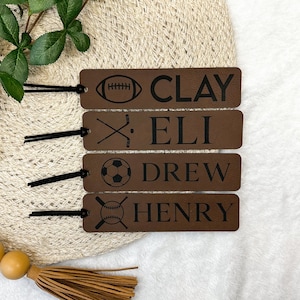 May include: Four brown leather bookmarks with black tassels. Each bookmark features a different sport icon and a name: Clay, Eli, Drew, and Henry.