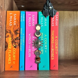 May include: A selection of fantasy novels by Sarah J. Maas, featuring titles such as "Silver Flames," "Frost and Starlight," "Wings and Ruin," "Mist and Fury," and "Thorns and Roses." A decorative bookmark and a small black figurine are also present.