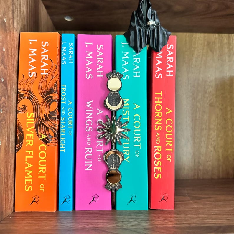 May include: A selection of fantasy novels by Sarah J. Maas, featuring titles such as "Silver Flames," "Frost and Starlight," "Wings and Ruin," "Mist and Fury," and "Thorns and Roses." A decorative bookmark and a small black figurine are also present.
