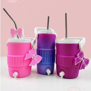 May include: Three colourful beverage coolers in pink, purple, and magenta, each with a white handle and spout. The coolers have a bow-shaped charm and a metal straw. The Luna Beam Creations logo is visible.