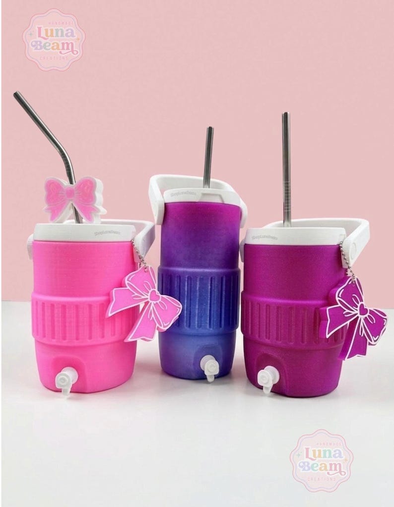 May include: Three colourful beverage coolers in pink, purple, and magenta, each with a white handle and spout. The coolers have a bow-shaped charm and a metal straw. The Luna Beam Creations logo is visible.