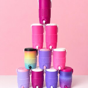 May include: A collection of brightly coloured beverage coolers in shades of pink, purple, and a rainbow gradient. Each cooler has a white lid, a spigot, and a handle. The coolers are arranged against a pink background.