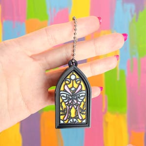 May include: A black stained glass window-shaped charm with a butterfly design. The charm has a yellow background with pink and white butterfly details. It hangs from a silver ball chain.