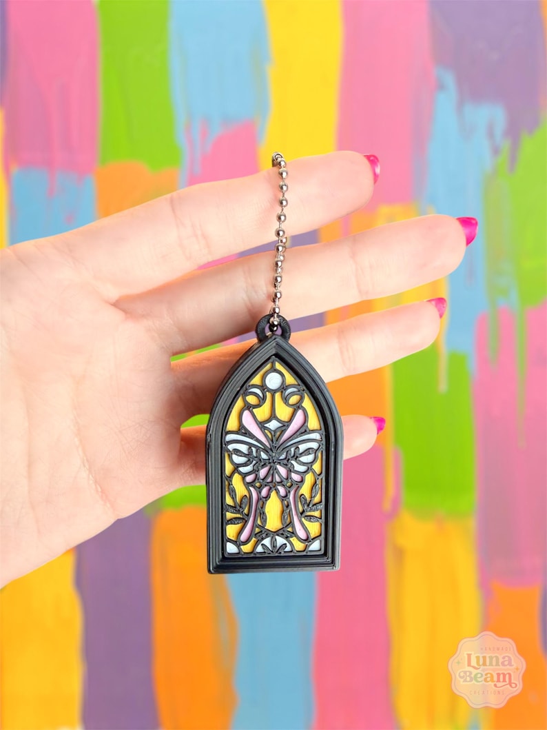 May include: A black stained glass window-shaped charm with a butterfly design. The charm has a yellow background with pink and white butterfly details. It hangs from a silver ball chain.
