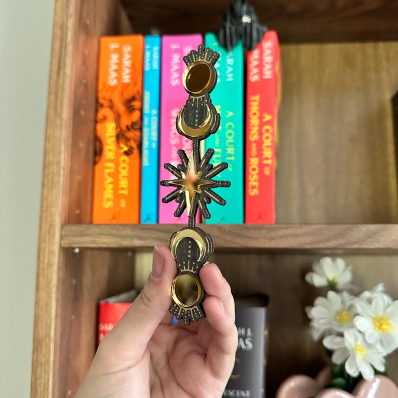 May include: A celestial-themed bookmark with gold star, moon, and crown details. The bookmark is held in front of a bookshelf with colourful book spines.