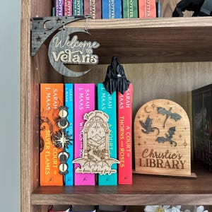 May include: A wooden bookshelf displaying a collection of books with vibrant covers. The shelf is adorned with wooden decorations, including a sign that reads "Welcome to Velaris." A small, black figure sits atop the books, adding a decorative touch.