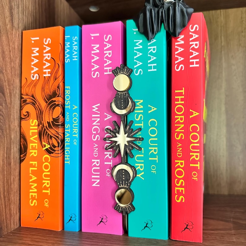 May include: A selection of books from the "A Court of Thorns and Roses" series by Sarah J. Maas, featuring vibrant covers. A black and gold bookmark with celestial motifs sits between the books. The books are displayed on a wooden shelf.