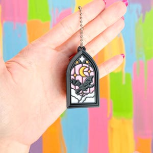 May include: A black and white stained glass-style keychain featuring a bat, moon, and stars design. The keychain has a silver chain and a black frame. The background is a colorful mix of pink, yellow, blue, and green.