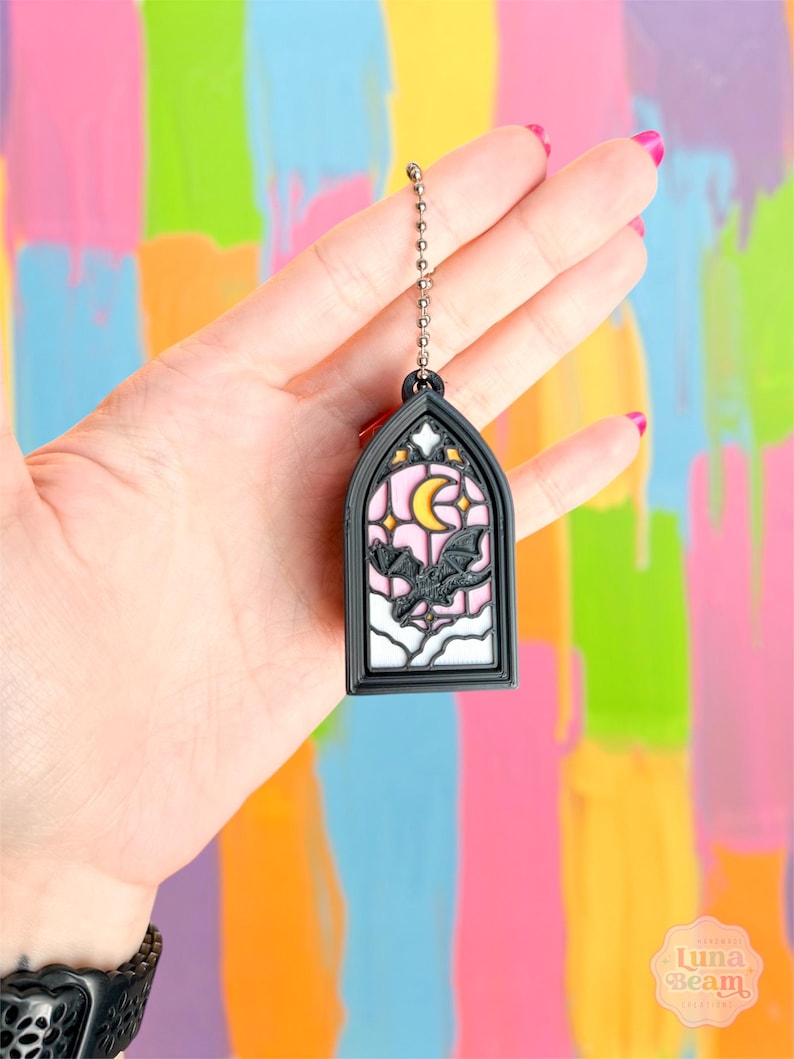 May include: A black and white stained glass-style keychain featuring a bat, moon, and stars design. The keychain has a silver chain and a black frame. The background is a colorful mix of pink, yellow, blue, and green.