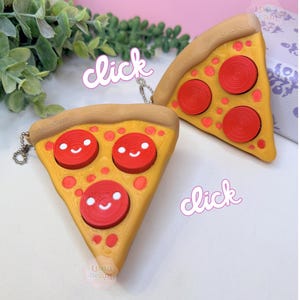 May include: Two pizza slice keychains with smiling pepperoni faces. The crust is a light brown, the cheese is yellow, and the pepperoni is red. The word "click" is written in a pink font.
