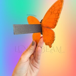 May include: A decorative butterfly bottle stopper with orange wings and a dark grey stem. The butterfly is held in a hand against a colourful gradient background. The text "LUNA BEAM" is visible.