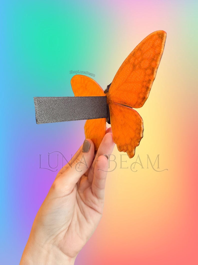 May include: A decorative butterfly bottle stopper with orange wings and a dark grey stem. The butterfly is held in a hand against a colourful gradient background. The text "LUNA BEAM" is visible.