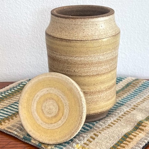 May include: A cylindrical ceramic canister with a speckled beige and tan glaze featuring horizontal bands. The canister has a matching circular lid with concentric yellow and cream rings, showcasing a textured, earthy finish throughout the piece.