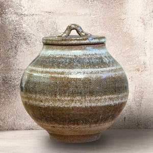 May include: A round ceramic jar with a lid, featuring a mottled glaze in shades of brown, tan, and white. The vessel has a wide, bulbous body that tapers toward the base and a small, arched handle centered on the top of the circular lid.