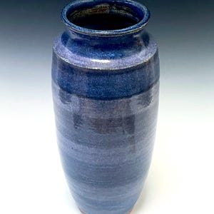 May include: A tall, handmade ceramic vase with a blue and purple glaze. The vase has a cylindrical shape with a slightly flared rim. The color transitions from a dark blue at the top to a lighter blue and purple towards the base. The base is a natural brown color.