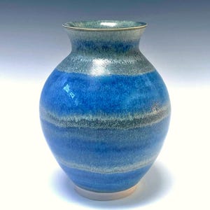 May include: A handcrafted ceramic vase with a rounded body and a flared rim. The vase features horizontal bands of blue, gray, and white, creating a layered effect. The base is a light beige color. This decorative vase is suitable for home decor.