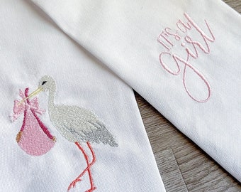 May include: White fabric sash with pink embroidery of a stork carrying a baby in a pink bundle. The sash has the text "It's a Girl" embroidered in pink.