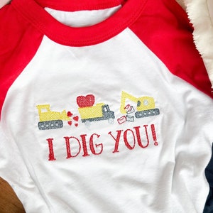 May include: White and red baseball tee shirt with a yellow construction vehicle graphic and the text "I DIG YOU!"