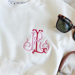 May include: A white sweatshirt with a pink and red embroidered monogram "R" on the front. The sweatshirt is laying on a white surface with a pair of brown sunglasses, a pair of blue jeans, and a brown and white handbag.