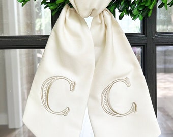 May include: A white satin bow with the letter "C" embroidered on each side, tied around a green boxwood wreath.