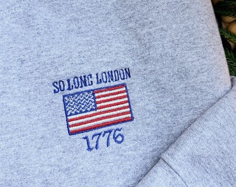 May include: Gray sweatshirt with embroidered text "So Long London" and an American flag with the year "1776" underneath.