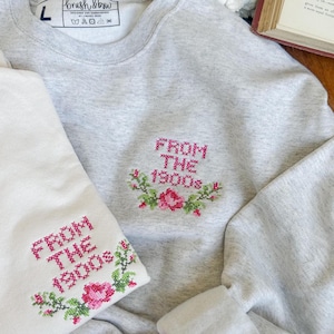 Könnte beinhalten: Ein graues Sweatshirt mit einer weißen Stickerei, die "From The 1900s" mit einem Blumenmuster sagt. Das Sweatshirt ist auf einer weißen Oberfläche mit einem Paar rosa Socken mit einem weißen Herzdesign gefaltet.